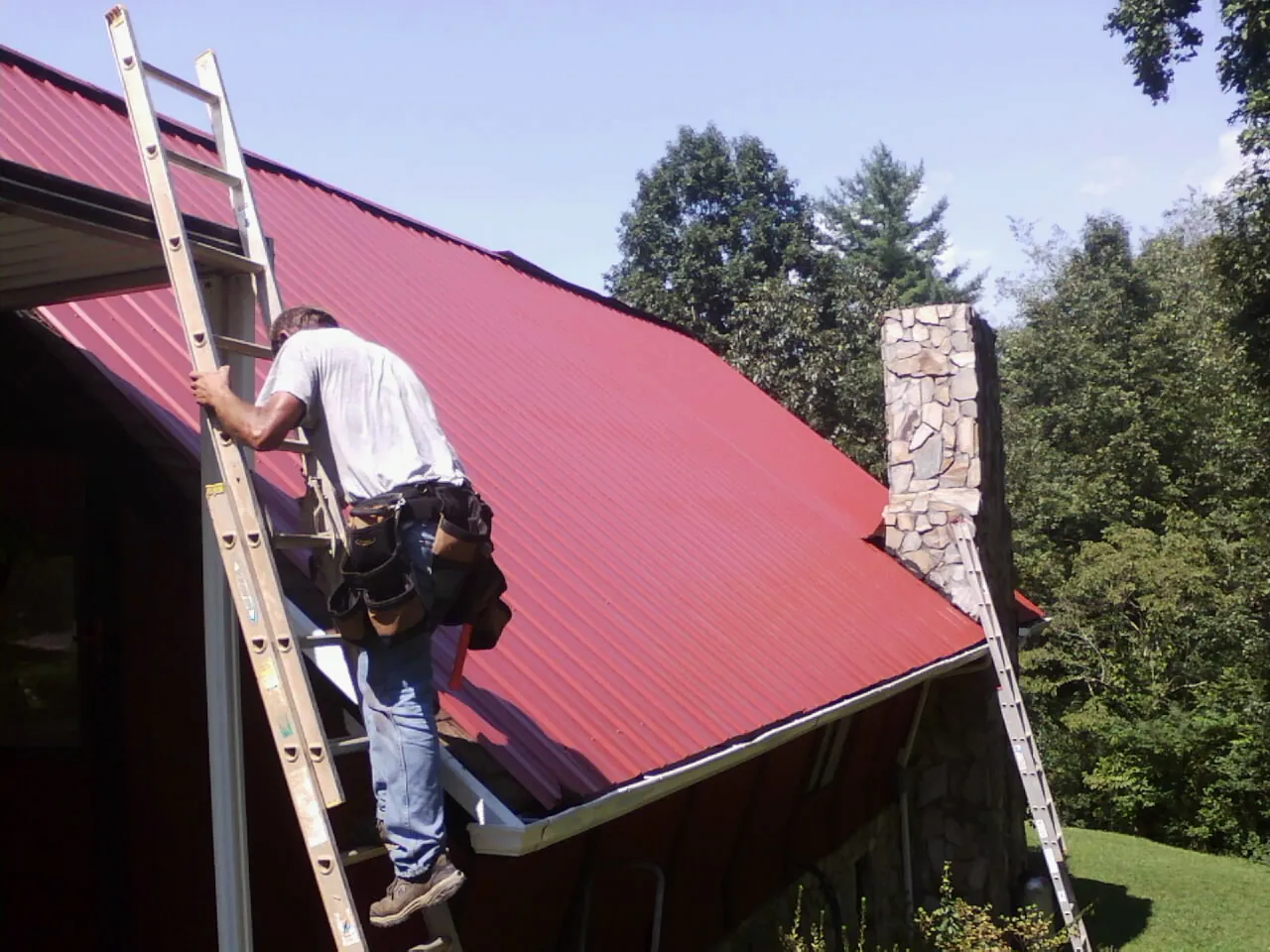Roofer on a ladder beside a red metal roof and stone chimney — Soffit & Fascia Repair by Roofing Brothers in Watsontown